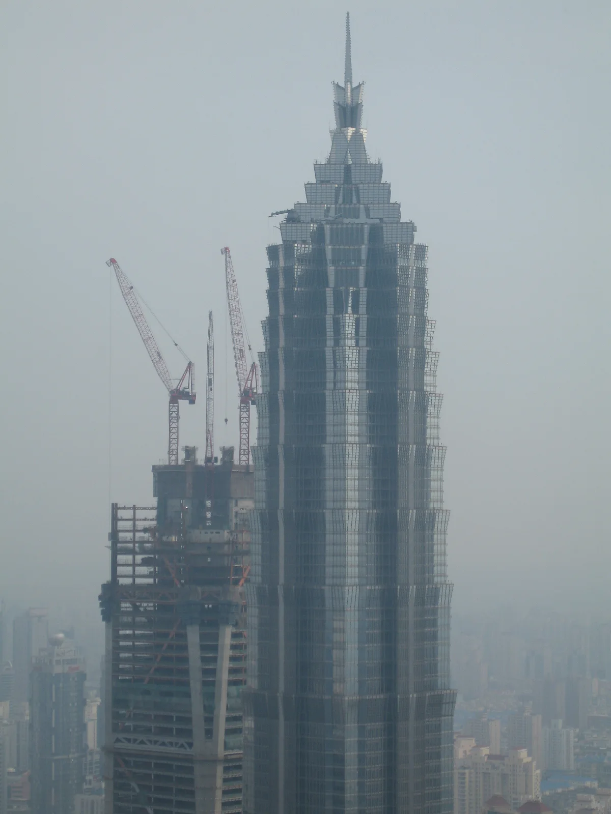 A stepped skyscraper with a narrow spire stands beside an unfinished building and crane in a hazy elevated view.