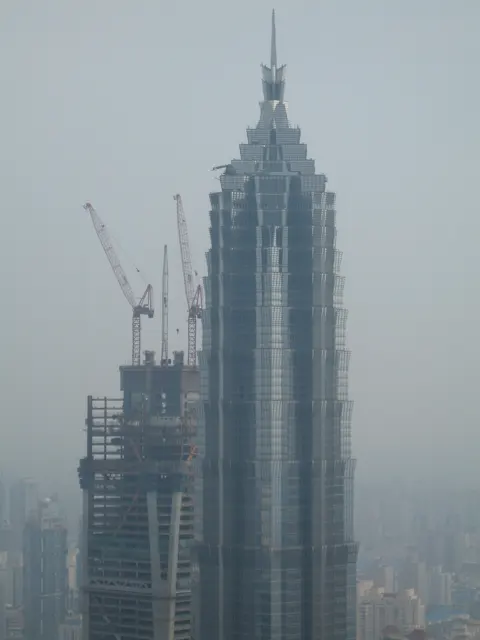 A stepped skyscraper with a narrow spire stands beside an unfinished building and crane in a hazy elevated view.