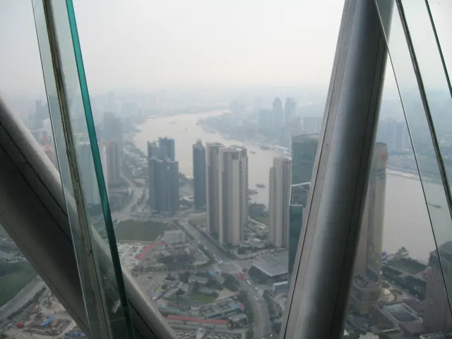 Sloping glass panels and large structural supports frame an elevated view of apartment blocks and streets.