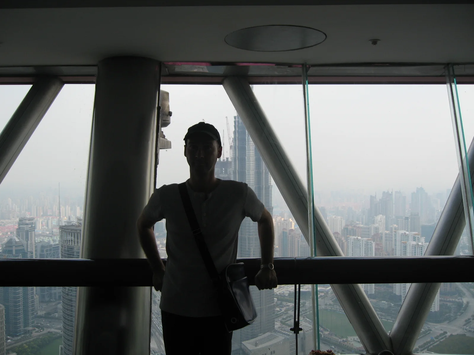 A person stands in silhouette beside broad observation windows and diagonal supports high above the city.