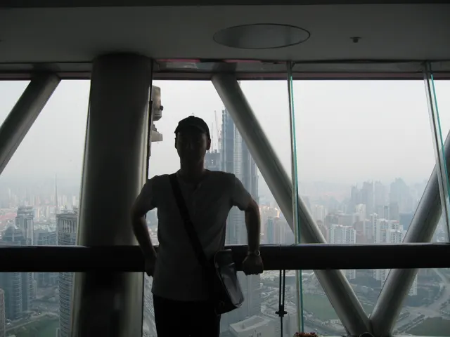 A person stands in silhouette beside broad observation windows and diagonal supports high above the city.