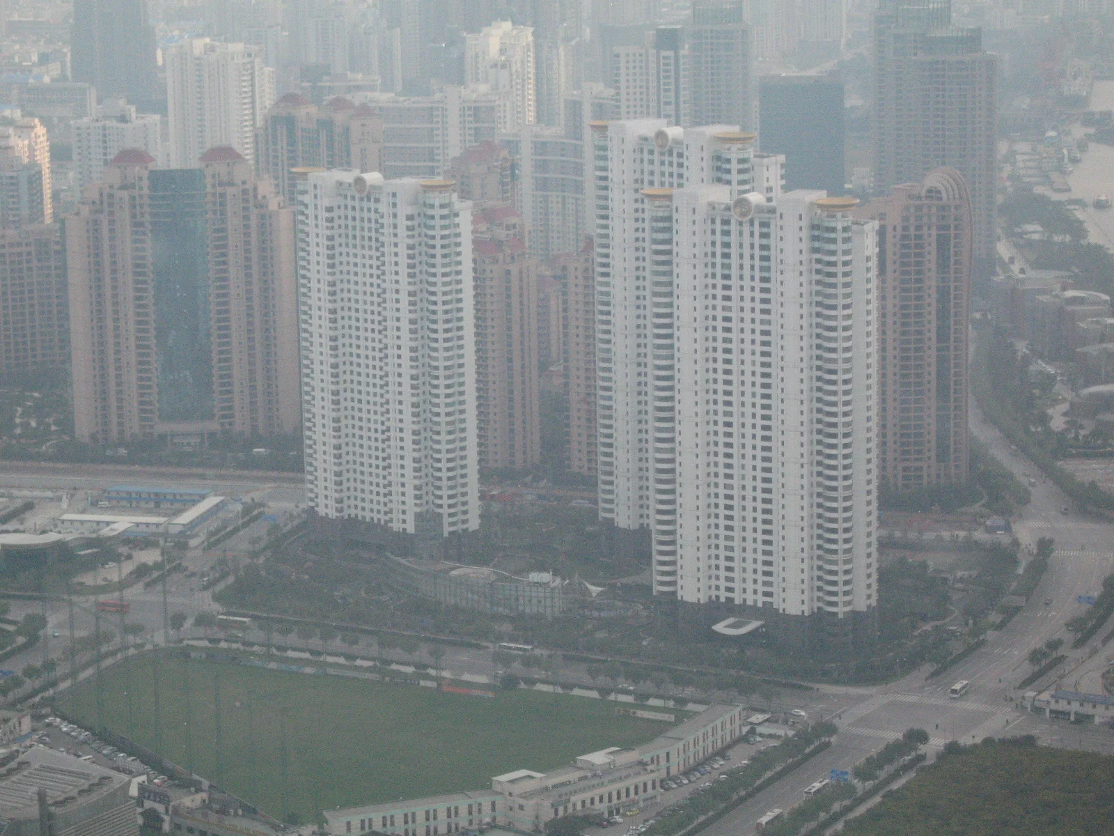 Groups of tall apartment buildings surround open ground and roads in a hazy overhead city view.