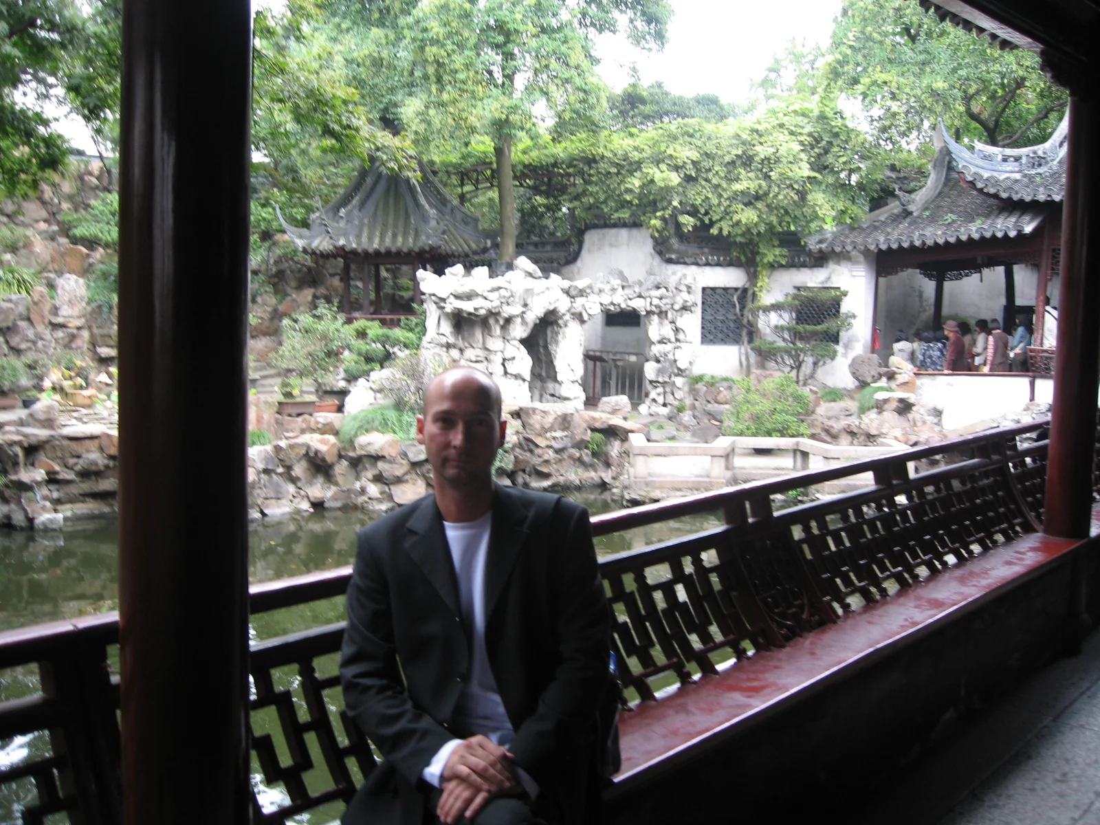 A person in a dark jacket sits beside a covered gallery railing overlooking rocks, plants and tiled garden pavilions.