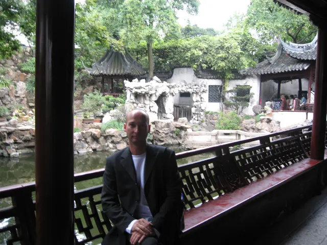 A person in a dark jacket sits beside a covered gallery railing overlooking rocks, plants and tiled garden pavilions.