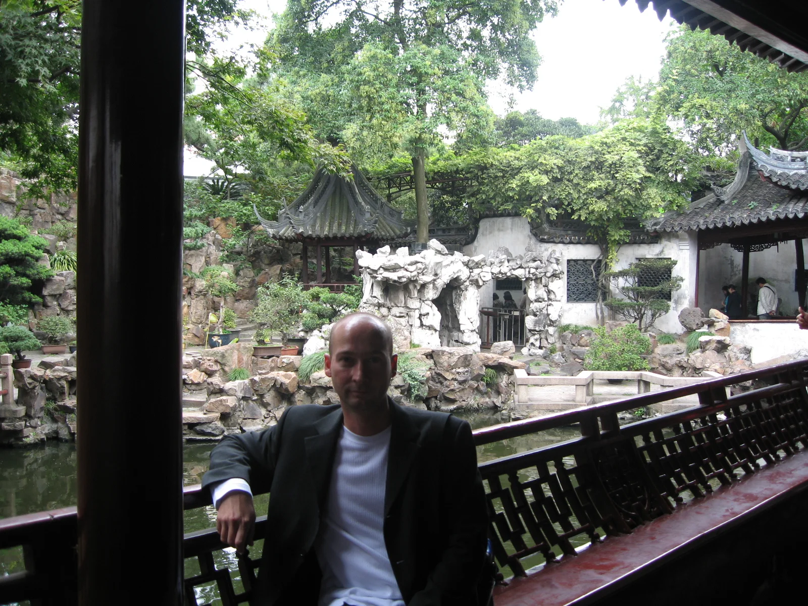 A person sits by a wooden railing under a covered walkway with a rock garden and white walls beyond.