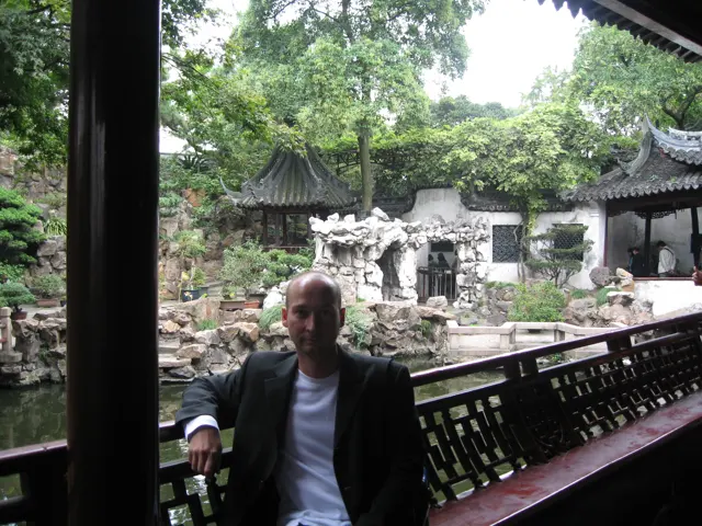 A person sits by a wooden railing under a covered walkway with a rock garden and white walls beyond.