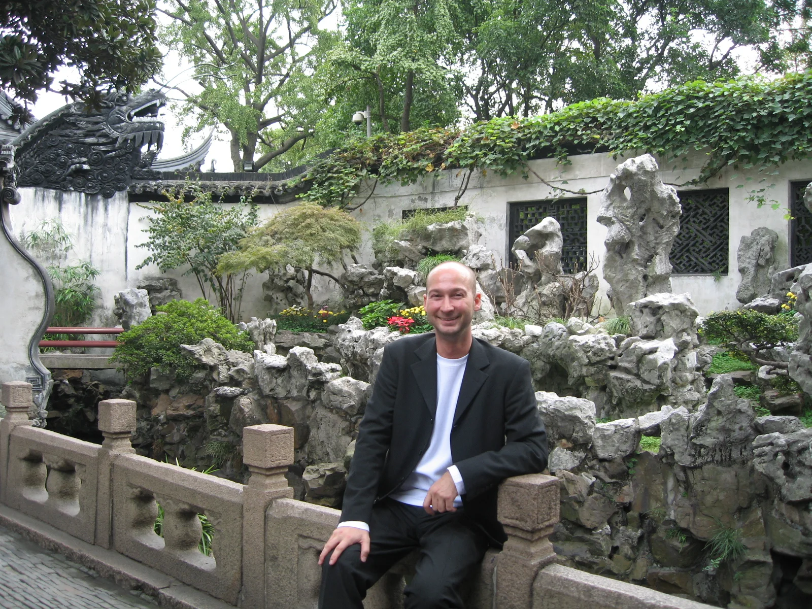 A person in a dark jacket sits on a carved stone railing with rocks, greenery and a white wall behind.