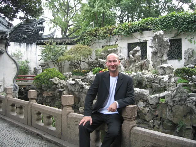 A person rests an arm on a stone balustrade beside a rock garden and white tiled-roof walls.