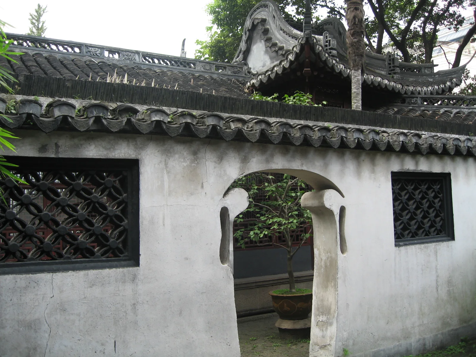 A curved doorway opens through a white wall with dark lattice windows and a tiled roof, with garden structures behind.