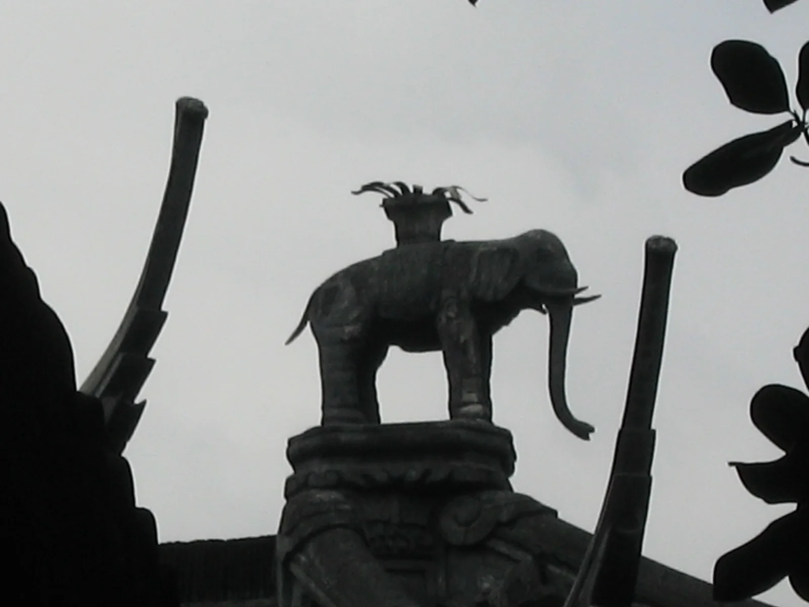 A small elephant sculpture with an object on its back stands in silhouette on a decorative base against a pale sky.