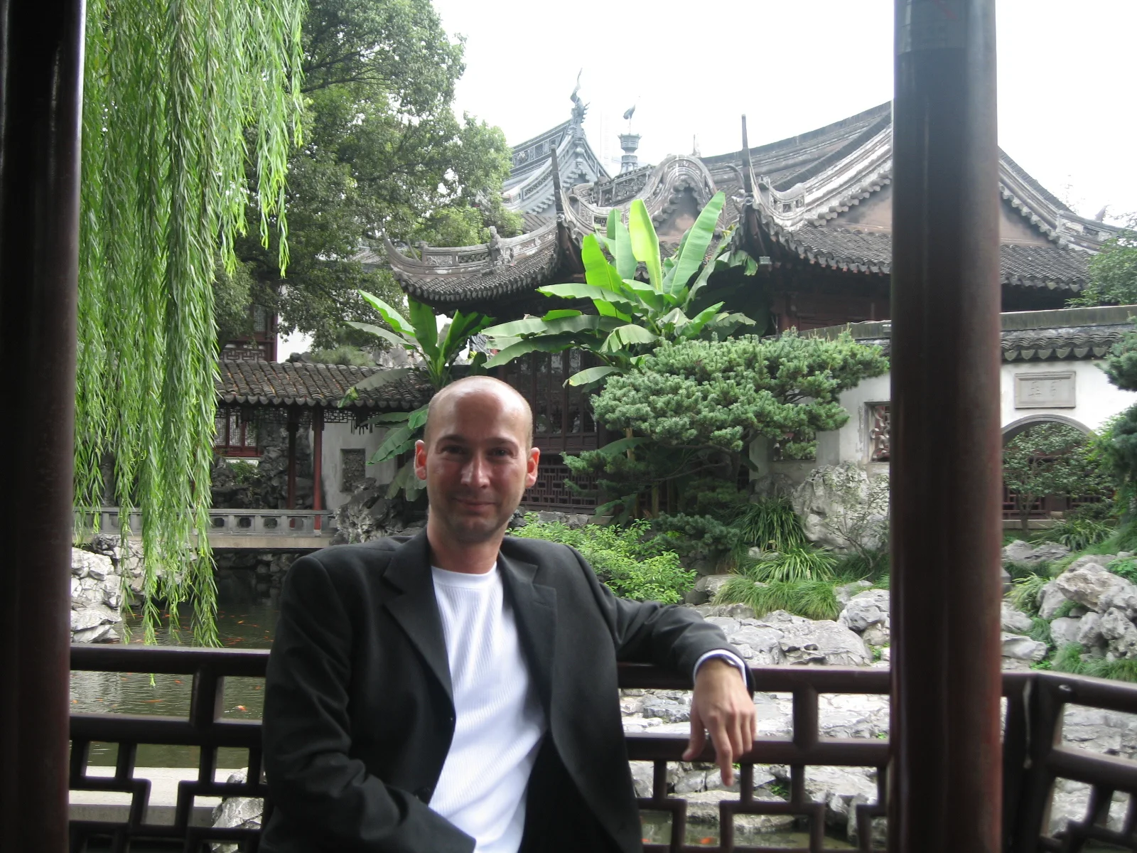 A person in a dark jacket sits beside a wooden garden railing with hanging greenery and tiled roofs behind.