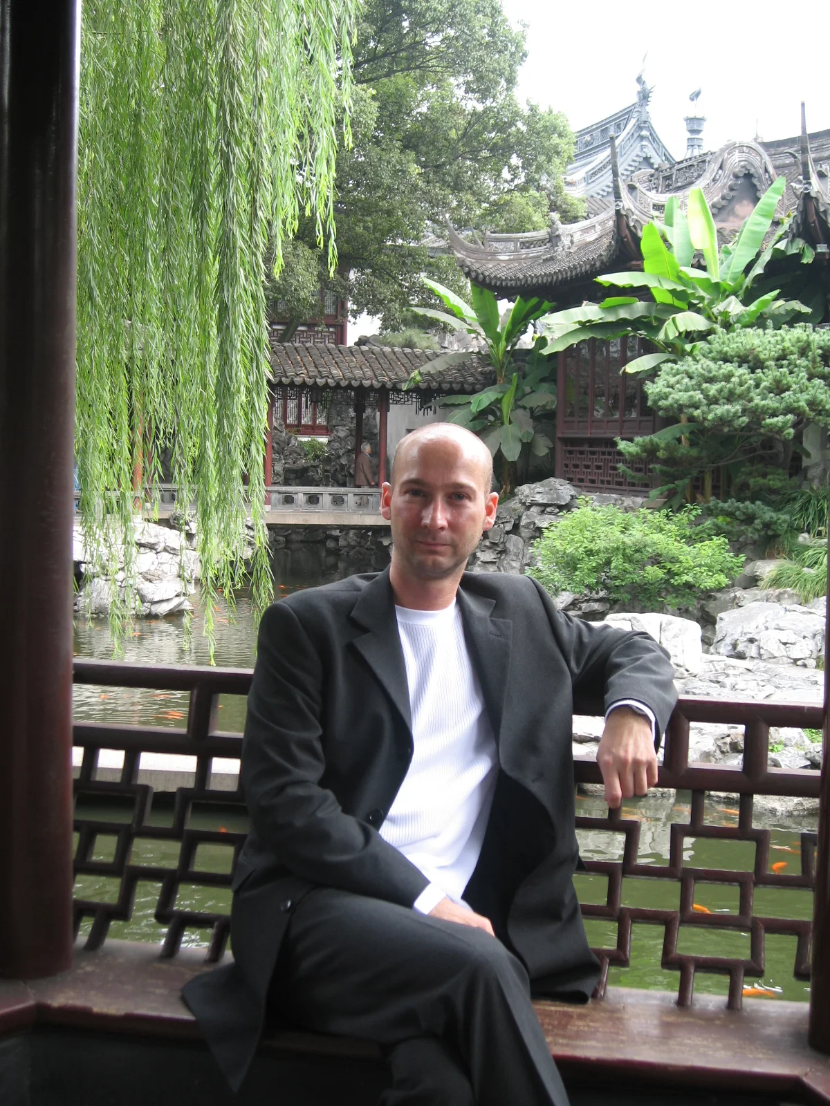 A seated person rests an arm on a railing beneath a covered gallery overlooking rocks, foliage and curved roofs.