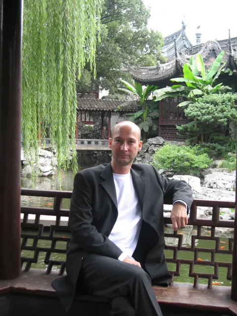 A seated person rests an arm on a railing beneath a covered gallery overlooking rocks, foliage and curved roofs.