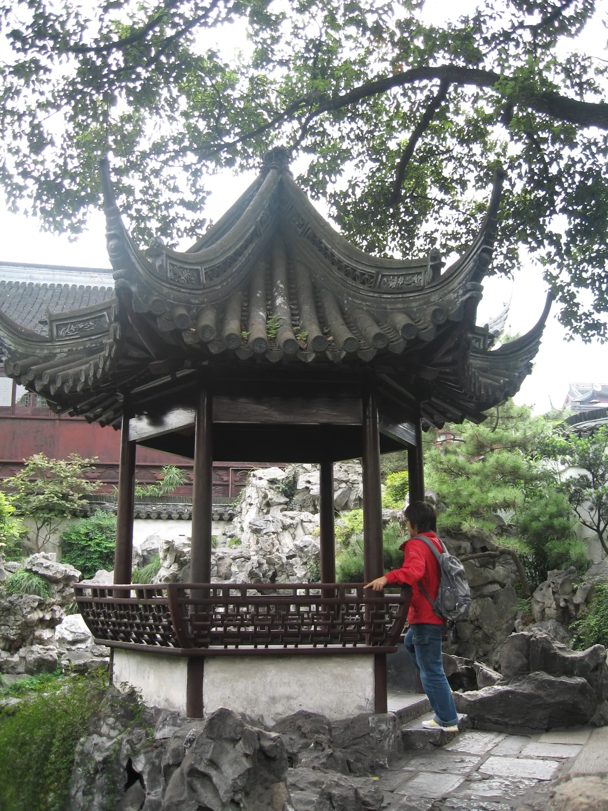 A small open pavilion with an upturned tiled roof stands among rocks and trees, with a person in red nearby.