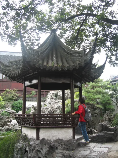 A small open pavilion with an upturned tiled roof stands among rocks and trees, with a person in red nearby.