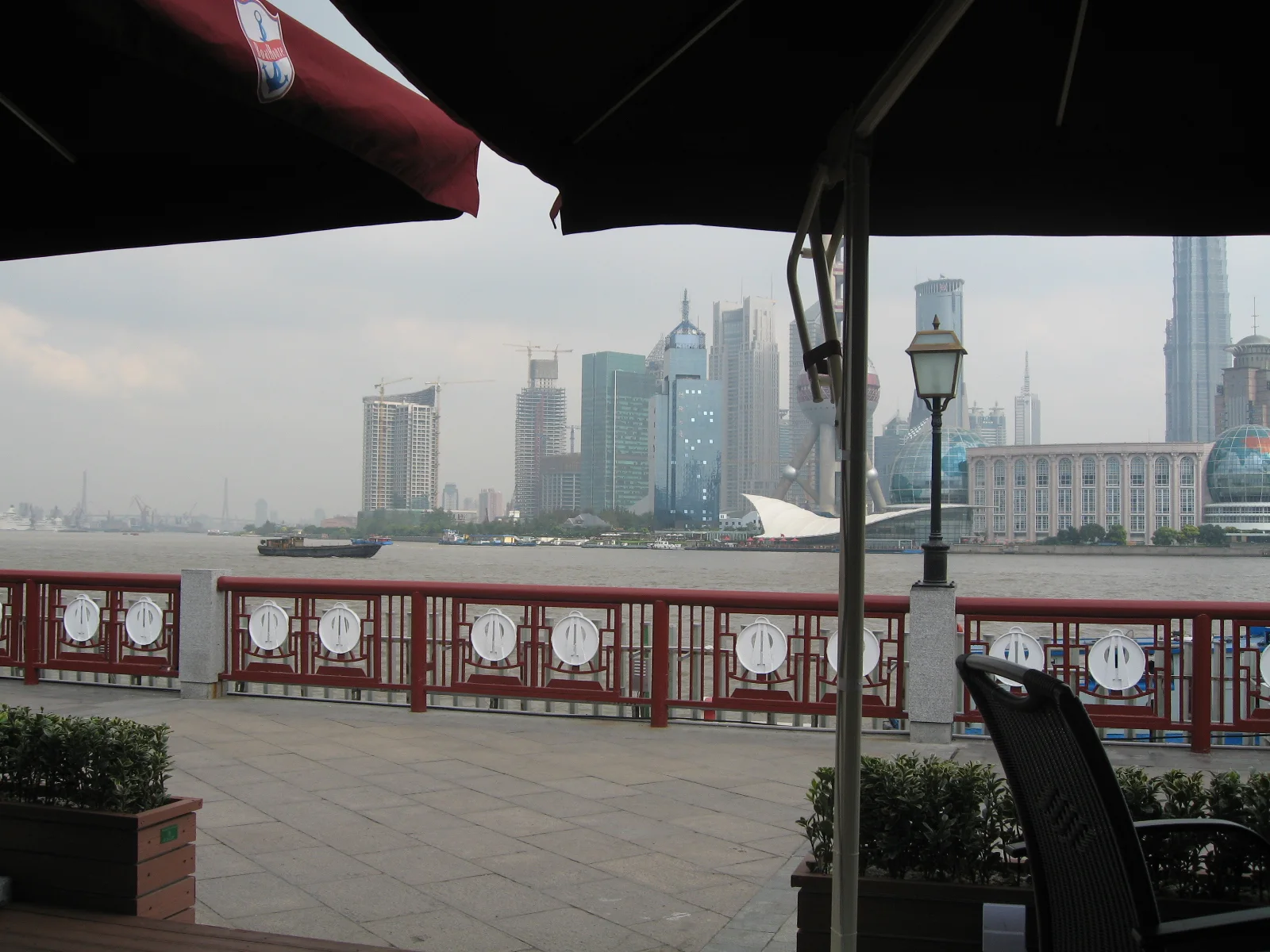 Dark terrace umbrellas frame a red riverside railing and a hazy skyline across the water.