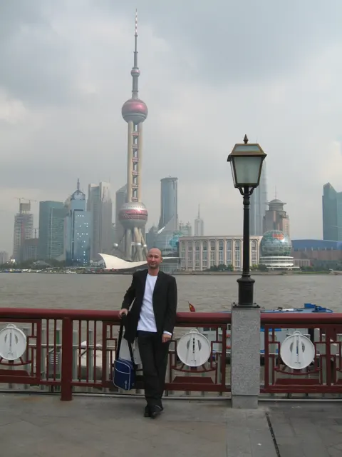 A person stands beside a decorative red railing and lamp post with a spherical-section tower across the river.