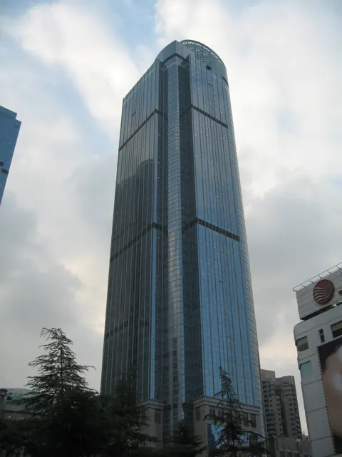 A tall blue-glass building with a rounded top rises above trees against a partly cloudy sky.
