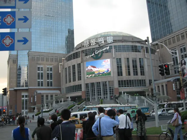Pedestrians gather at a crossing before a curved shopping-centre facade with a large screen and vehicles passing in front.