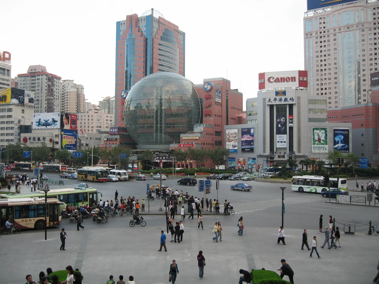 An elevated view looks across a busy paved square towards a large glass sphere and buildings covered with signs.