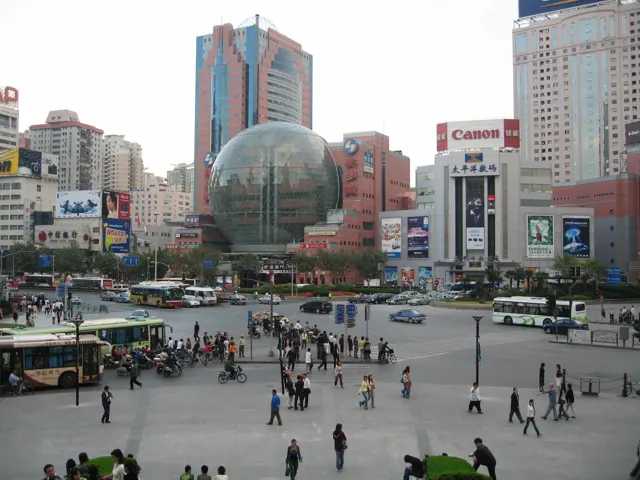 An elevated view looks across a busy paved square towards a large glass sphere and buildings covered with signs.