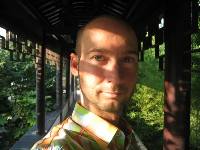 A close portrait of a person in a patterned shirt is crossed by bands of sunlight beneath a shaded garden corridor.