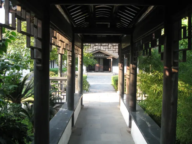 A narrow covered garden passage leads between wooden columns and geometric lattice panels towards a bright courtyard.