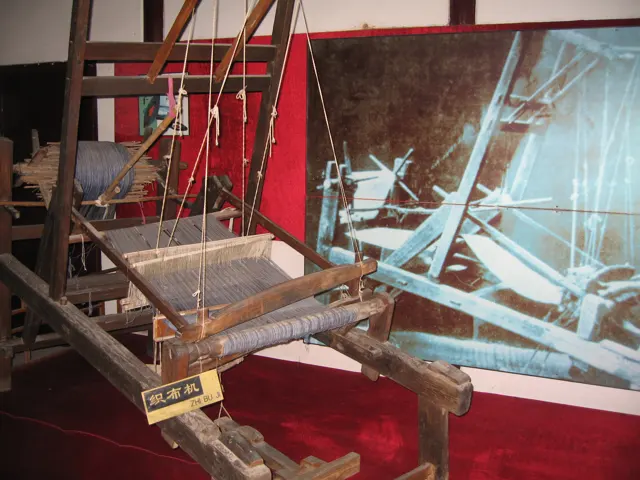 A wooden loom with suspended threads is displayed indoors against red panels and an explanatory image.