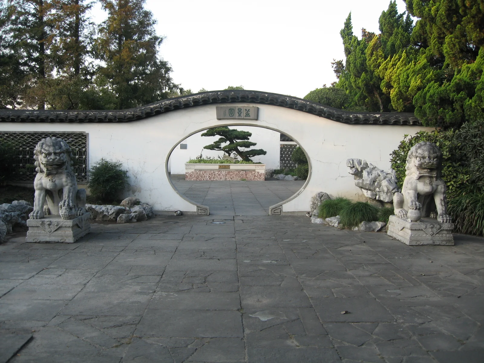 A circular opening in a white garden wall frames a shaped tree, with stone guardian figures at either side.