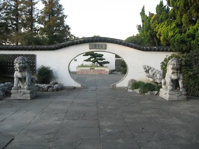 A circular opening in a white garden wall frames a shaped tree, with stone guardian figures at either side.