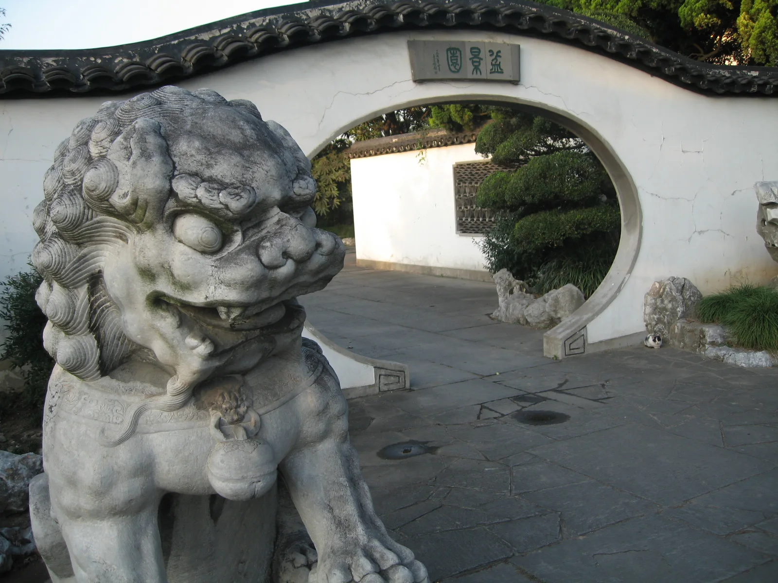 A carved stone lion stands in the foreground beside a circular opening through a white wall.