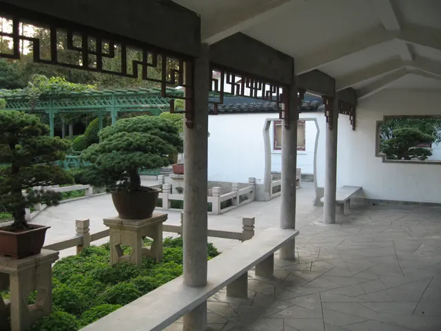 A covered gallery with pale columns borders a display of potted trees and stone railings.
