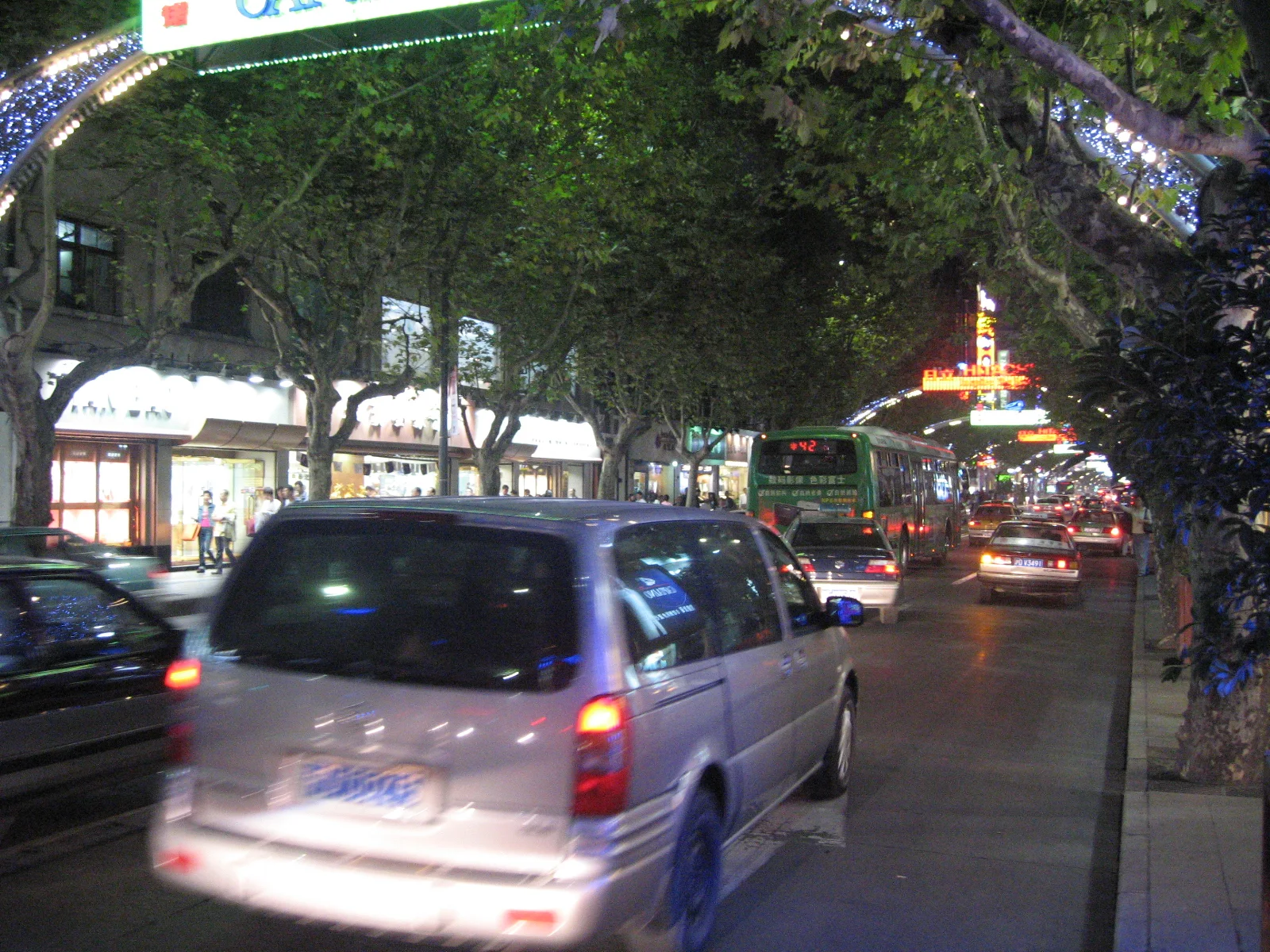 Cars pass beneath decorative lights spanning a tree-lined street at night, with a lit pavilion-like structure beyond.