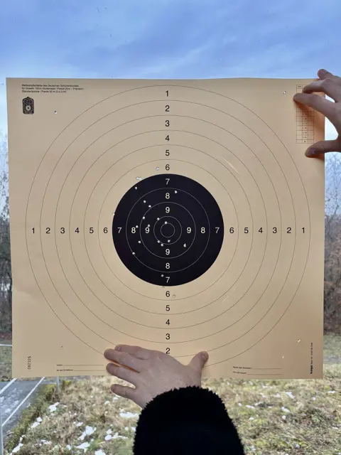 Hands hold up a square paper target outdoors, with a blue sky above and a dark sleeve below.