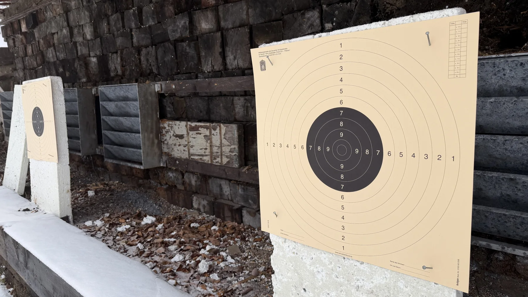 Two paper targets stand in front of a dark timber backstop, with the nearer target on the right above a pale support.