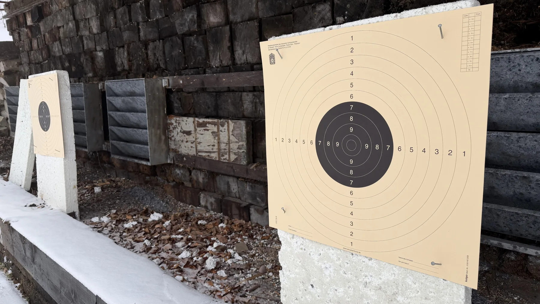 An angled view along two paper targets and pale supports beside a backstop of stacked dark timber.