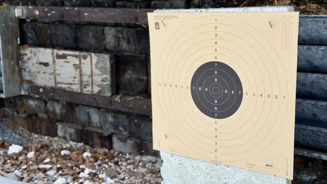 A cream target sheet with a black centre stands on a pale support in front of weathered timber and scattered debris.