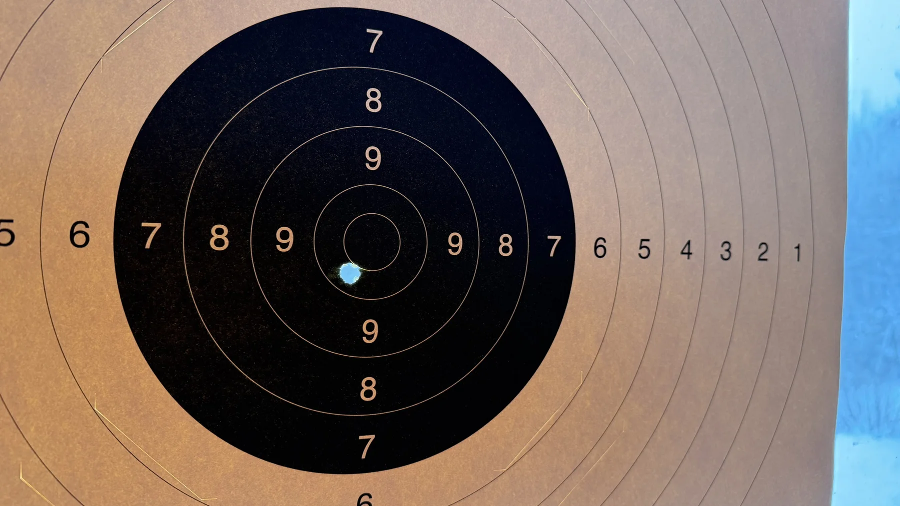 A bright opening appears below and to the left of the innermost circle on a black-and-cream paper target.