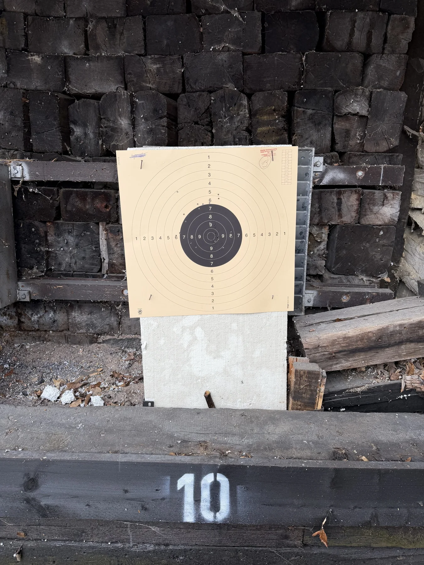A second portrait view of the target and its pale support, framed by stacked timber and a lower beam marked 10.