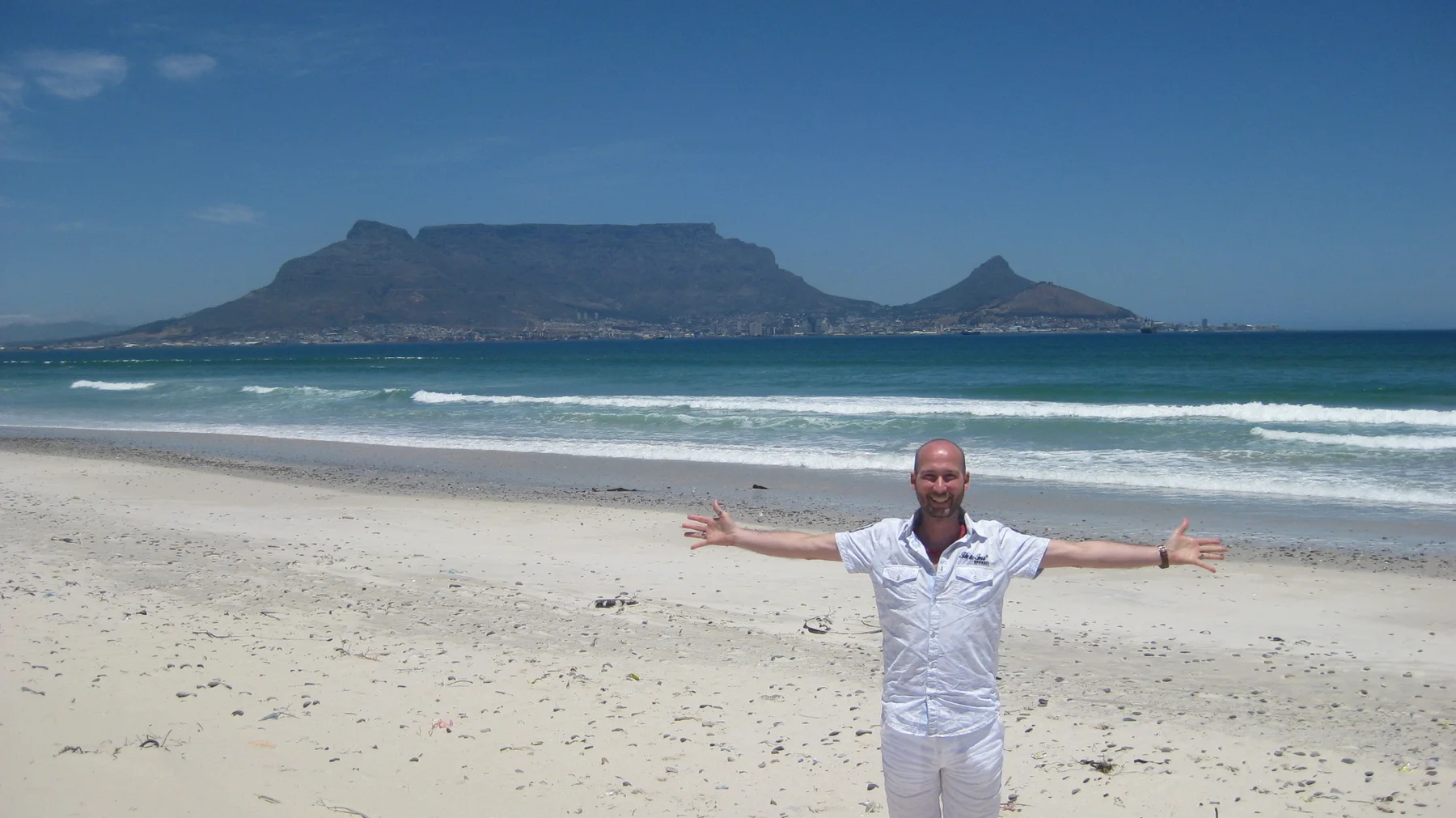 A person dressed in white stands with arms outstretched on a sandy beach, with a flat-topped mountain across the water.