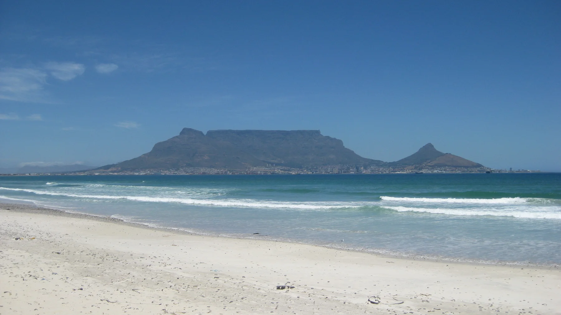 A broad, flat-topped mountain rises across turquoise water beyond a pale sandy beach.