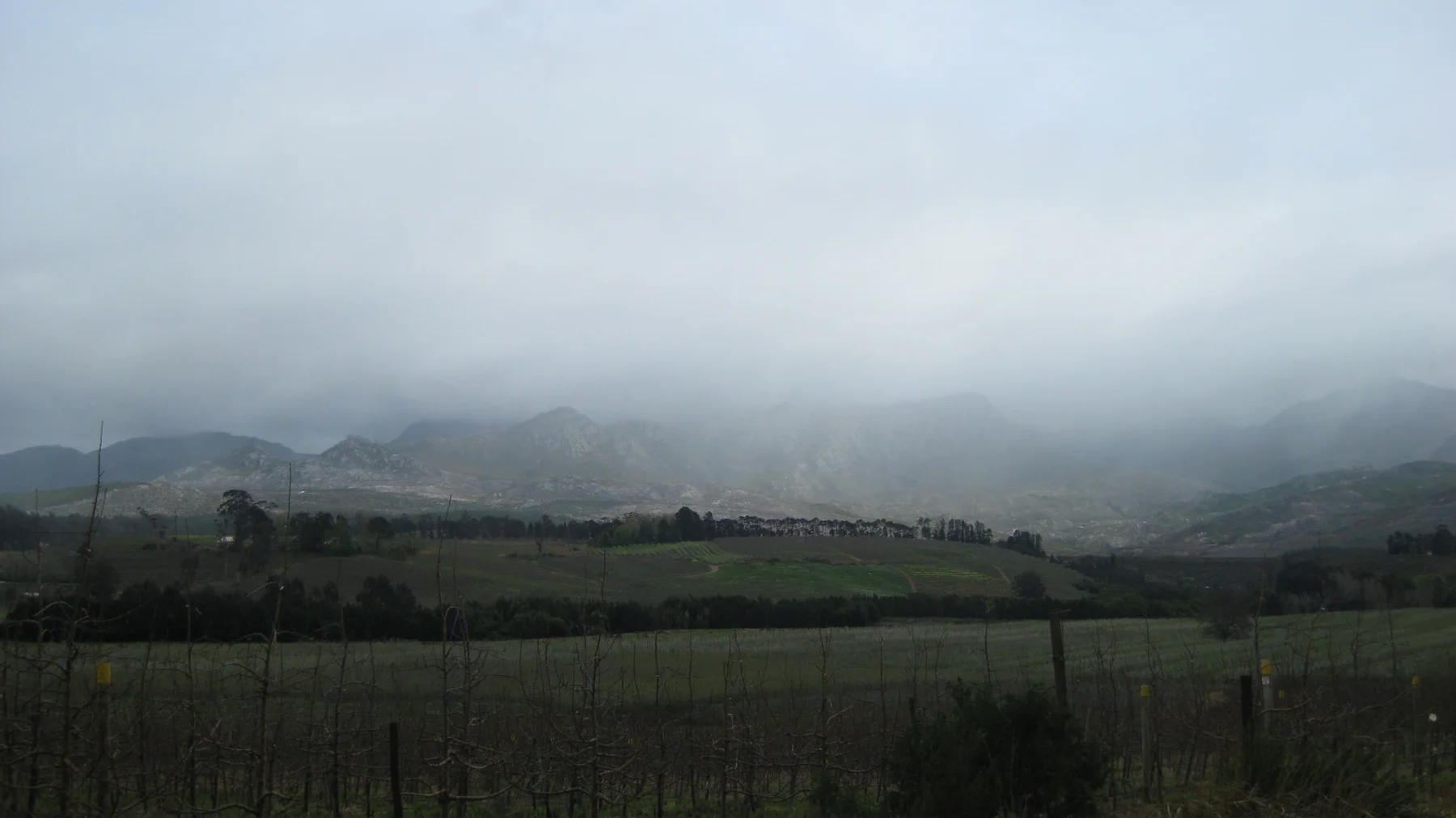 Low clouds obscure the mountains beyond fenced fields and dark green vegetation.