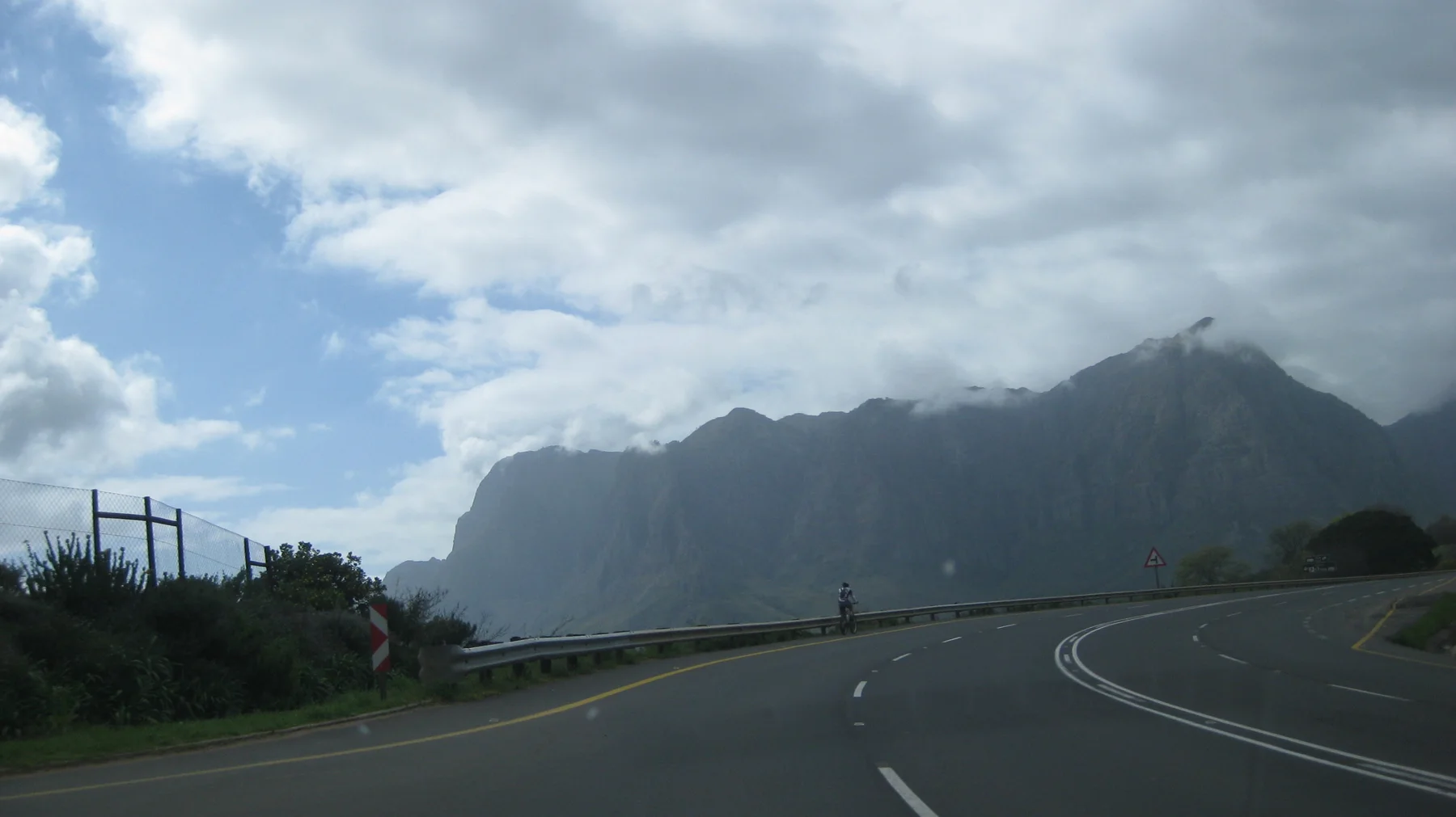 A paved road curves toward a steep mountain ridge partly wrapped in cloud.