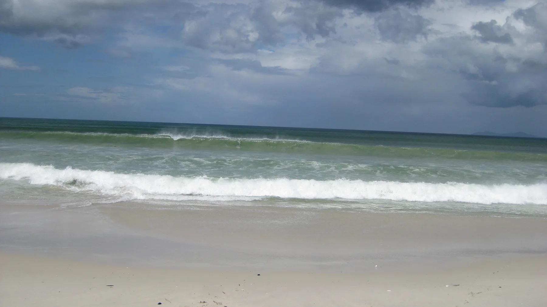 White waves roll onto a sandy beach beneath a sky filled with dark and bright clouds.