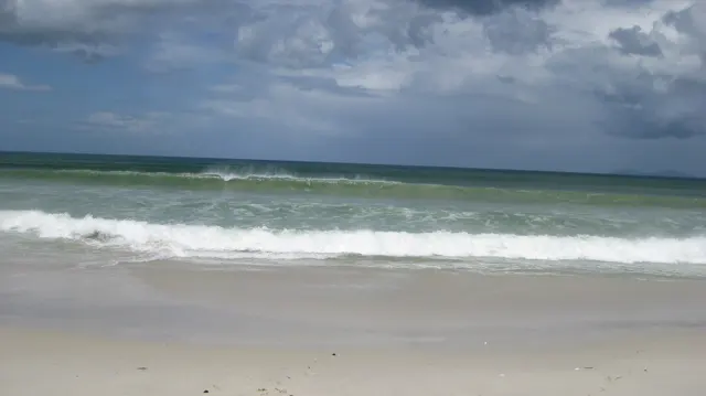 White waves roll onto a sandy beach beneath a sky filled with dark and bright clouds.