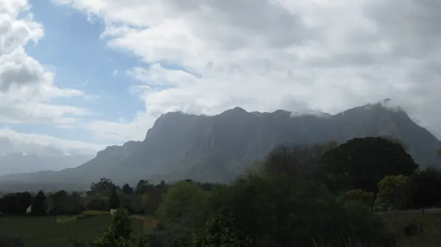 A rugged mountain wall rises above trees and green fields with cloud spilling over its crest.