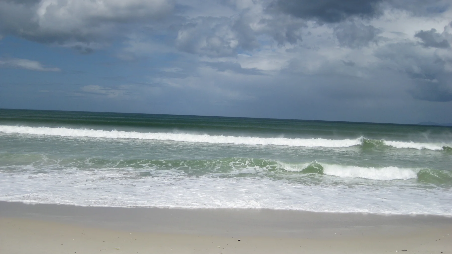Parallel lines of white surf cross green-blue water beneath a cloud-filled sky.