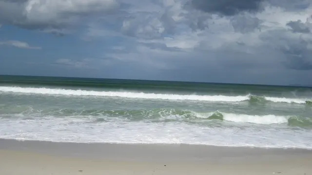 Parallel lines of white surf cross green-blue water beneath a cloud-filled sky.