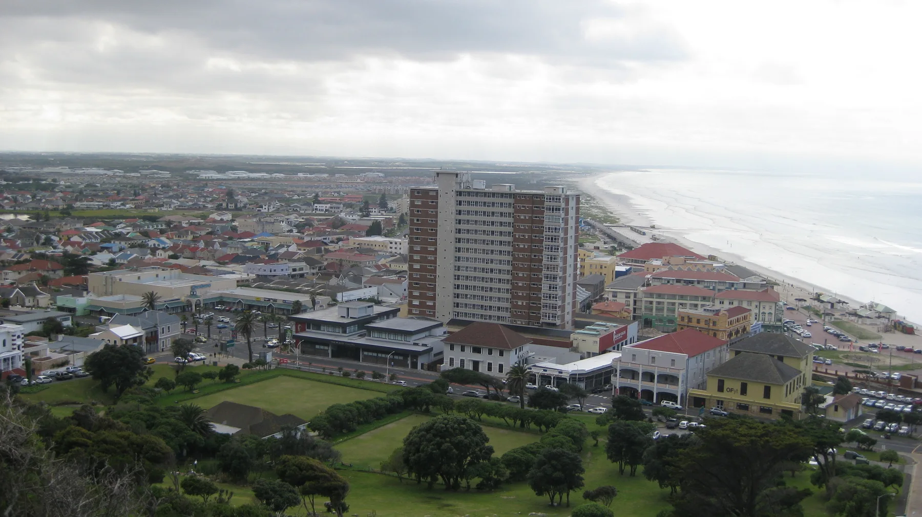 An elevated view looks across town buildings, a tall apartment block and a long sandy coastline.