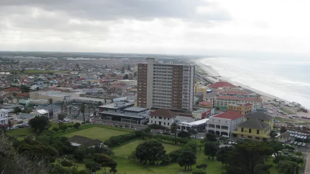 An elevated view looks across town buildings, a tall apartment block and a long sandy coastline.
